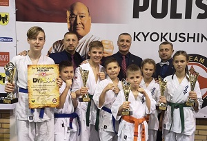 reprezentacja UKS Karate Kyokushin „MUSHIN” Szydłowiec razem z trenerem i sędziemy. Zawodnicy trzymają w dłoniach zwycięskie puchary.